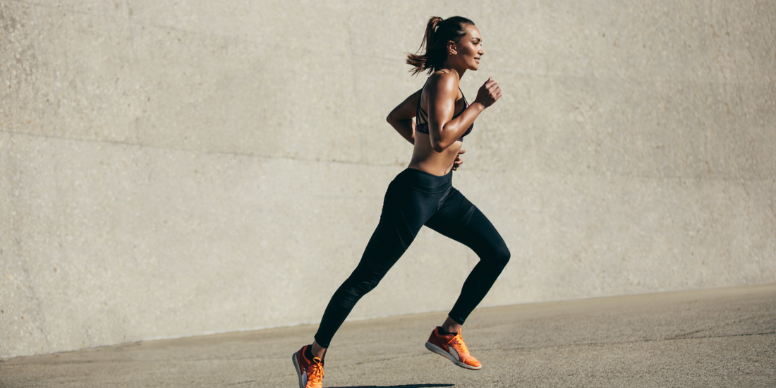 Junge sportliche Frau läuft dynamisch an einer Betonwand entlang, trägt schwarze Sportleggings und orange Laufschuhe bei Tageslicht.